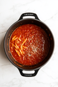 this image shows A pot filled with crushed tomatoes, vegetable broth, uncooked penne pasta, salt, and pepper, all combined to cook together in a one-pot pasta dish.