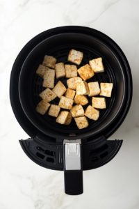 This image shows tofu strips being air-fried in an air fryer basket, turning golden brown and crispy.