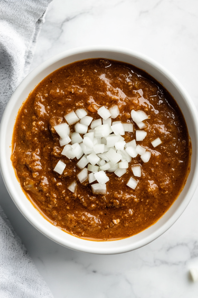 This image shows a bowl of Grandpa’s classic coney sauce, slow-cooked and topped with freshly chopped onions for a traditional touch.