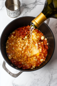 this image shows White wine being poured into the pot with sautéed tomato paste and spices, deglazing the pan and reducing for added depth of flavor in the pasta sauce.