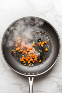 this image shows Chopped onions and carrots being sautéed in a hot pan, forming the flavorful base for the creamy Vegan Pasta Primavera sauce.