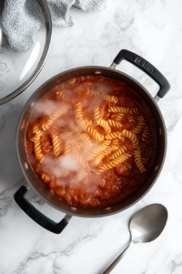 this image shows Penne pasta simmering in a rich tomato sauce, absorbing the flavors as it cooks to a tender consistency.