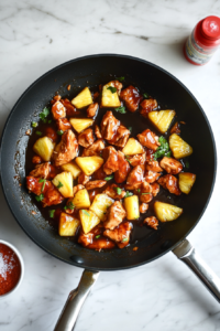 This image shows teriyaki sauce and sriracha being poured into the skillet with chicken and pineapple, creating a rich, tangy, and spicy sauce for the dish.