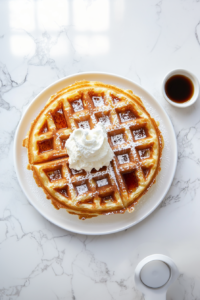 This image shows a crispy golden waffle served on a plate, topped with powdered sugar, maple syrup, and a dollop of fresh cream, making a delicious and indulgent breakfast.