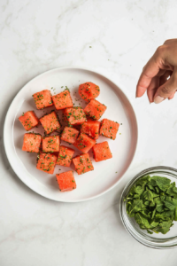 This image shows finely chopped fresh mint leaves being sprinkled over Tajín-seasoned watermelon slices, enhancing the dish with a refreshing herbal note.