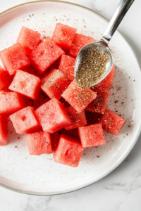 This image shows a hand sprinkling Tajín seasoning generously over juicy watermelon slices, adding a perfect balance of tangy, spicy, and salty flavors.