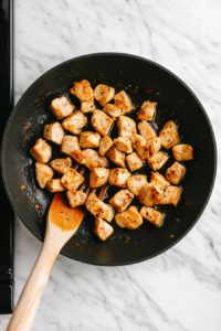 This image shows bite-sized chicken pieces sizzling in a pan, turning golden brown as they cook to perfection for a delicious teriyaki bowl.