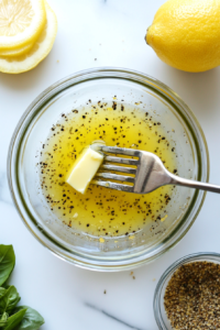 This image shows a small saucepan with melted butter, freshly squeezed lemon juice, and cracked black pepper being stirred together to create a rich, tangy, and buttery lemon pepper sauce.