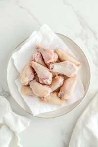 This image shows raw chicken wings being patted dry with a paper towel on a cutting board, ensuring they are properly prepped before seasoning and baking for the perfect crispy texture.