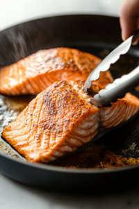 This image shows a salmon fillet sizzling in a hot pan, developing a flavorful golden-brown crust on the first side.
