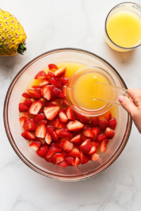 This image shows a top-down view of pineapple juice and lemon juice being poured into a large glass punch bowl, creating a sweet and tangy base for the punch.