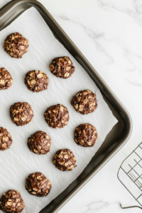 This image shows freshly scooped cookies resting on parchment paper as they cool and firm up. Their slightly rough texture, filled with visible oats and chocolate, captures the essence of homemade, no-bake goodness.