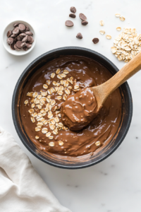 This image shows a thick and gooey chocolate mixture as hearty oats and fragrant vanilla extract are being stirred in, creating a rich and textured cookie dough. The wooden spoon smoothly incorporates all ingredients for even distribution.