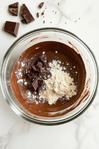 This image shows a clear glass bowl filled with flour, baking powder, and salt on a white marble countertop, ready to be combined.
