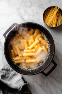 This image shows penne pasta being cooked in a large pot of salted boiling water until just shy of al dente.