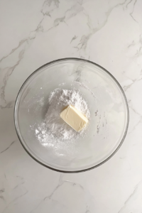 This image shows a glass mixing bowl with partially creamed butter, sugar, and salt on a white marble surface, with an electric mixer beside it.
