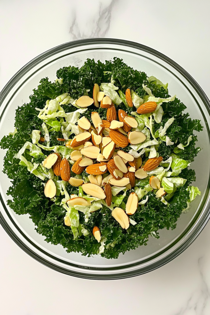 This image shows the finished Chick-fil-A kale salad in a glass bowl topped with roasted almond slices, set on a white marble countertop.