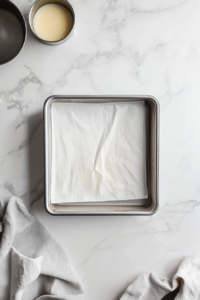 This image shows the oven being preheated to 325°F and an 8×8-inch baking pan being greased and lined with parchment paper, with extra overhang on the sides.