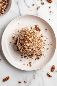 This image shows the chilled cheese ball being rolled in chopped pecans, creating a crunchy outer layer ready to be served.