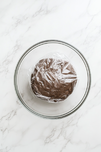 This image shows chocolate cookie dough covered with plastic wrap inside a clear glass bowl on a white marble countertop.