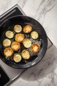 This image shows breaded zucchini slices frying in batches inside a black nonstick skillet over a white marble cooktop.