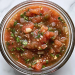 This image shows a top-down shot of vibrant homemade salsa served in a round clear glass bowl, placed on a clean white marble countertop. The salsa is chunky and colorful with visible bits of tomato, onion, cilantro, and jalapeño. The background is completely clean and minimal.
