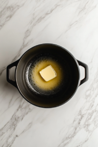 This image shows a large black soup pot on a white marble countertop with melted butter inside, ready to sauté vegetables.