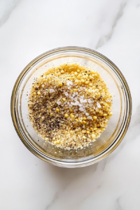 This image shows cornmeal, salt, and pepper mixed in a clear glass bowl placed on a clean white marble cooktop.
