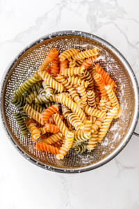 This image shows cooked rotini pasta being rinsed with cold water in a stainless steel colander over a clean white marble surface.