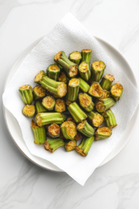 This image shows freshly fried golden okra draining on a paper towel-lined white plate on a white marble cooktop.