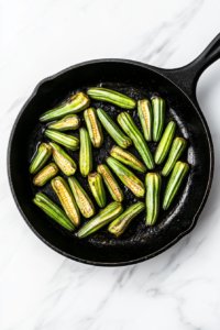 This image shows okra turning golden and crispy as it continues to fry in a black cast iron skillet on a white marble cooktop.