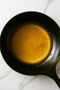 This image shows oil heating in a black cast iron skillet placed on a white marble cooktop with no background items.