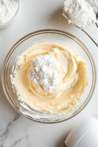 adding-powdered-sugar-to-frosting-in-clear-glass-bowl