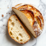 This image shows a rustic, golden sourdough loaf with a shallow slit on top, resting on white parchment paper placed over a white marble countertop. Clean, minimal, top-down shot.